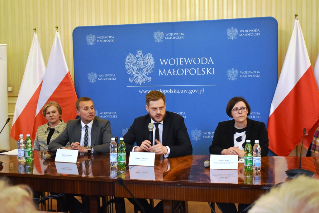 zdjęcie przedstawiające cztery siedzące osoby przy stole na tle ścianki Urzędu Wojewódzkiego